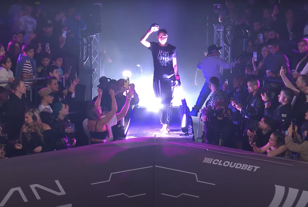 Aline Pereira walks out to her second fight with Karate Combat. In the picture a spot light shines on her black shirt with white letters reading, "Jesus Loves You."